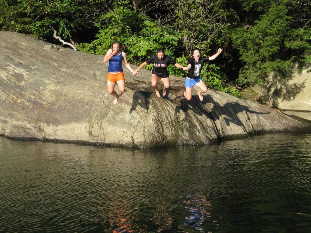Swimmers shoulder-deep in clear green water at Lareau Swim Hole with a large rock face and summer forest behind them.