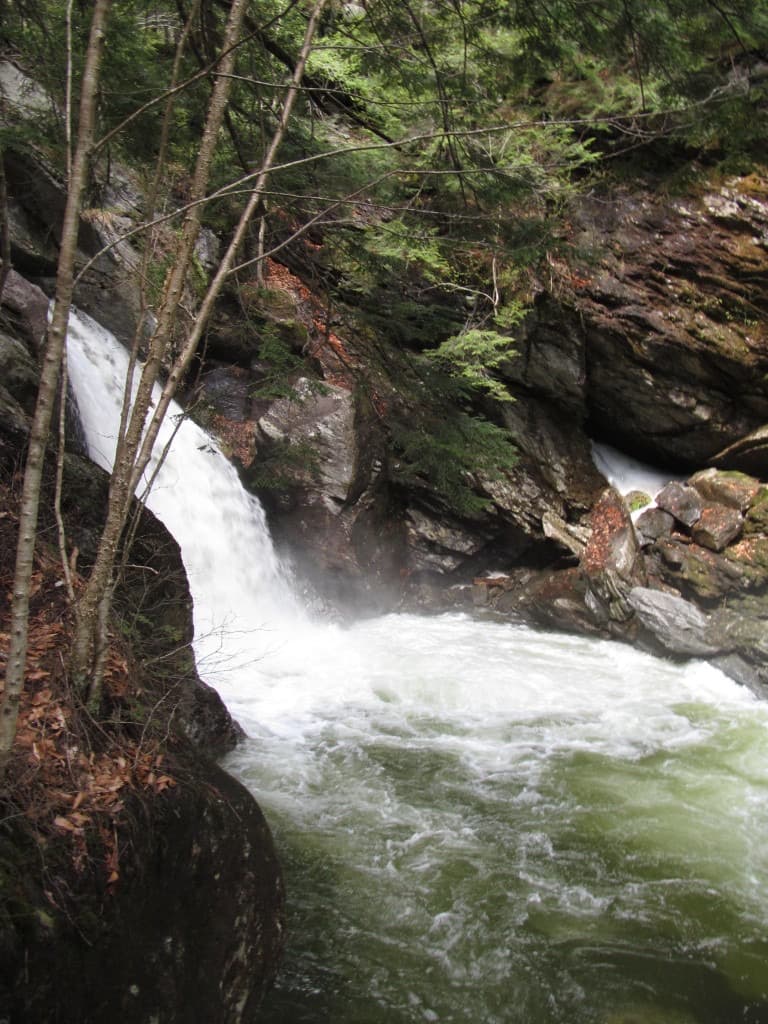 A waterfall at Bingham Falls pouring over jagged rock into a churning green pool, with evergreens and bare trees along the gorge rim.