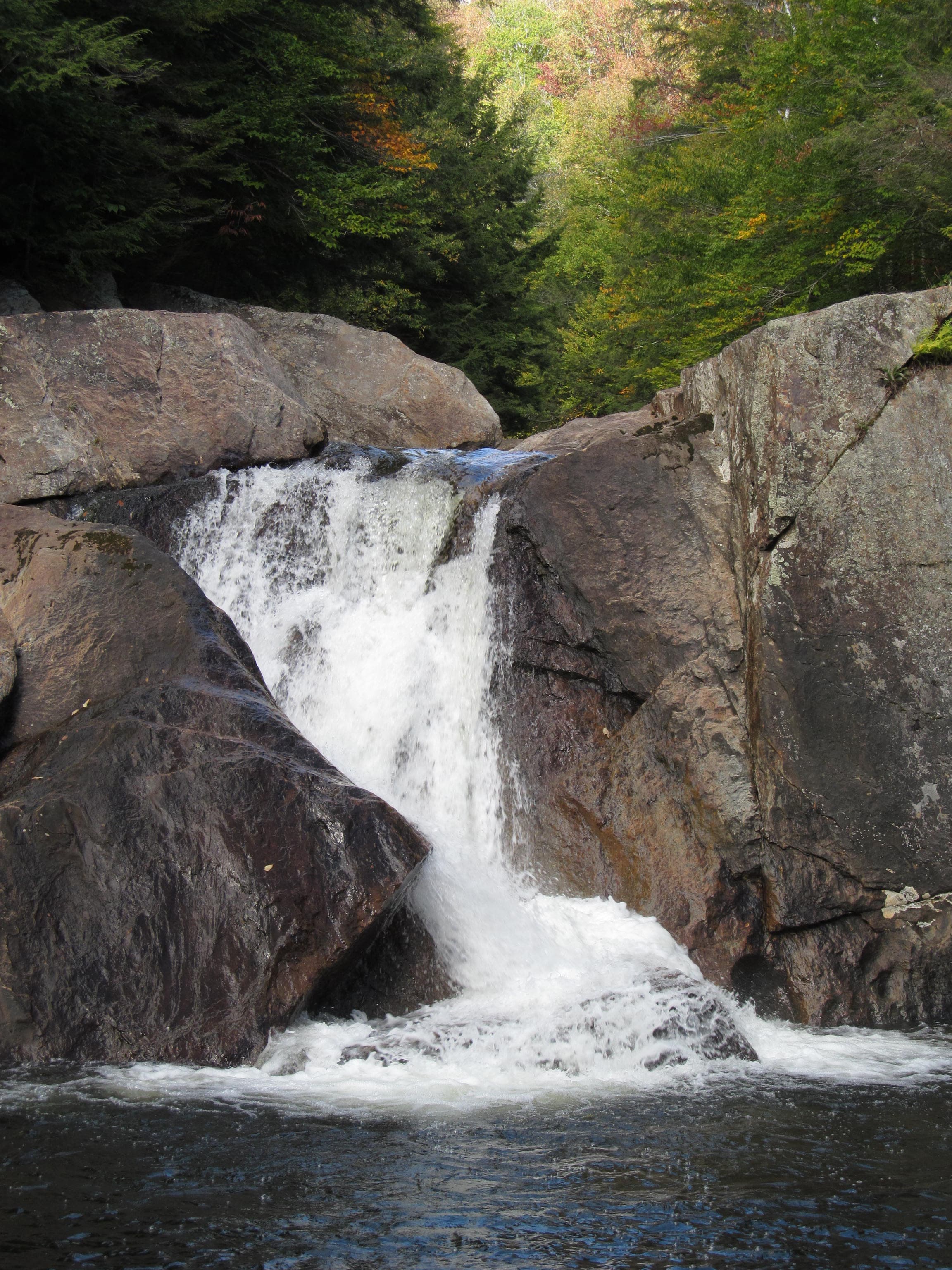 Waterfall plunging through a rock notch into a dark pool, mixed forest with early fall color at Buttermilk Falls, Ludlow, Vermont.
