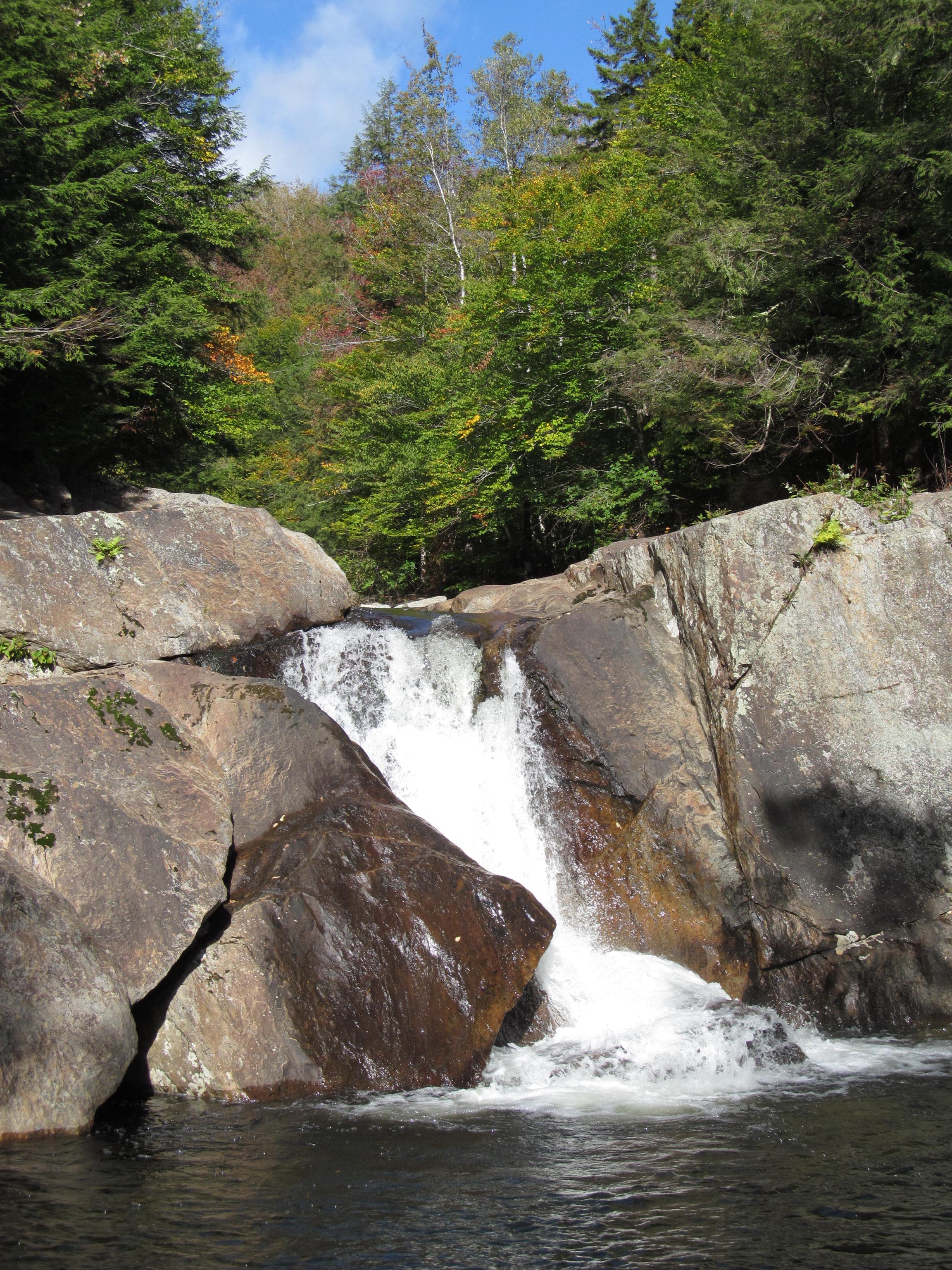 Whitewater dropping between two massive grey boulders into a pool, forested ridge behind Buttermilk Falls, Ludlow, Vermont.