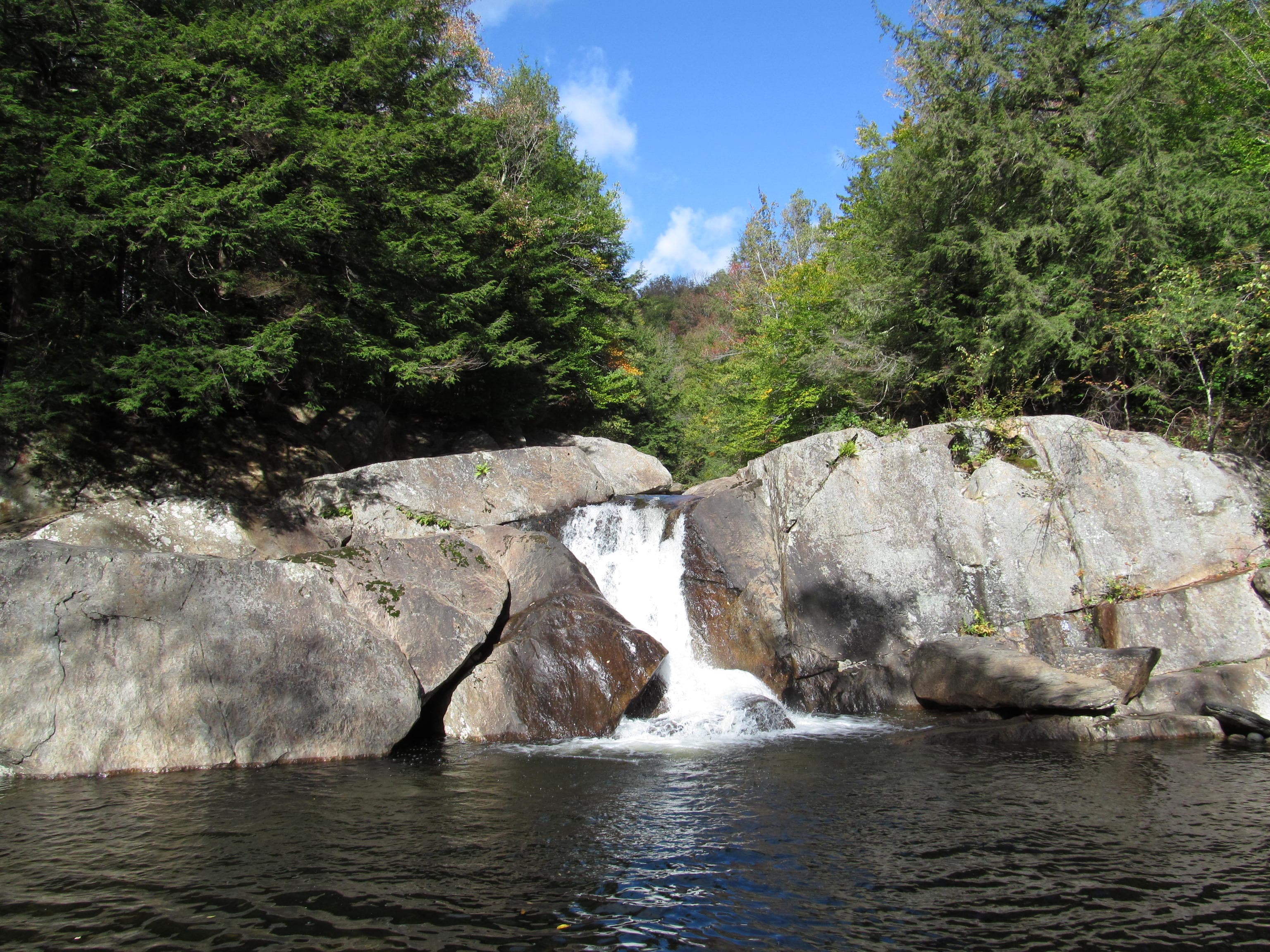 Wide waterfall flowing over smooth grey ledges into a dark pool at Buttermilk Falls in Ludlow, Vermont, with forested banks and blue sky above.