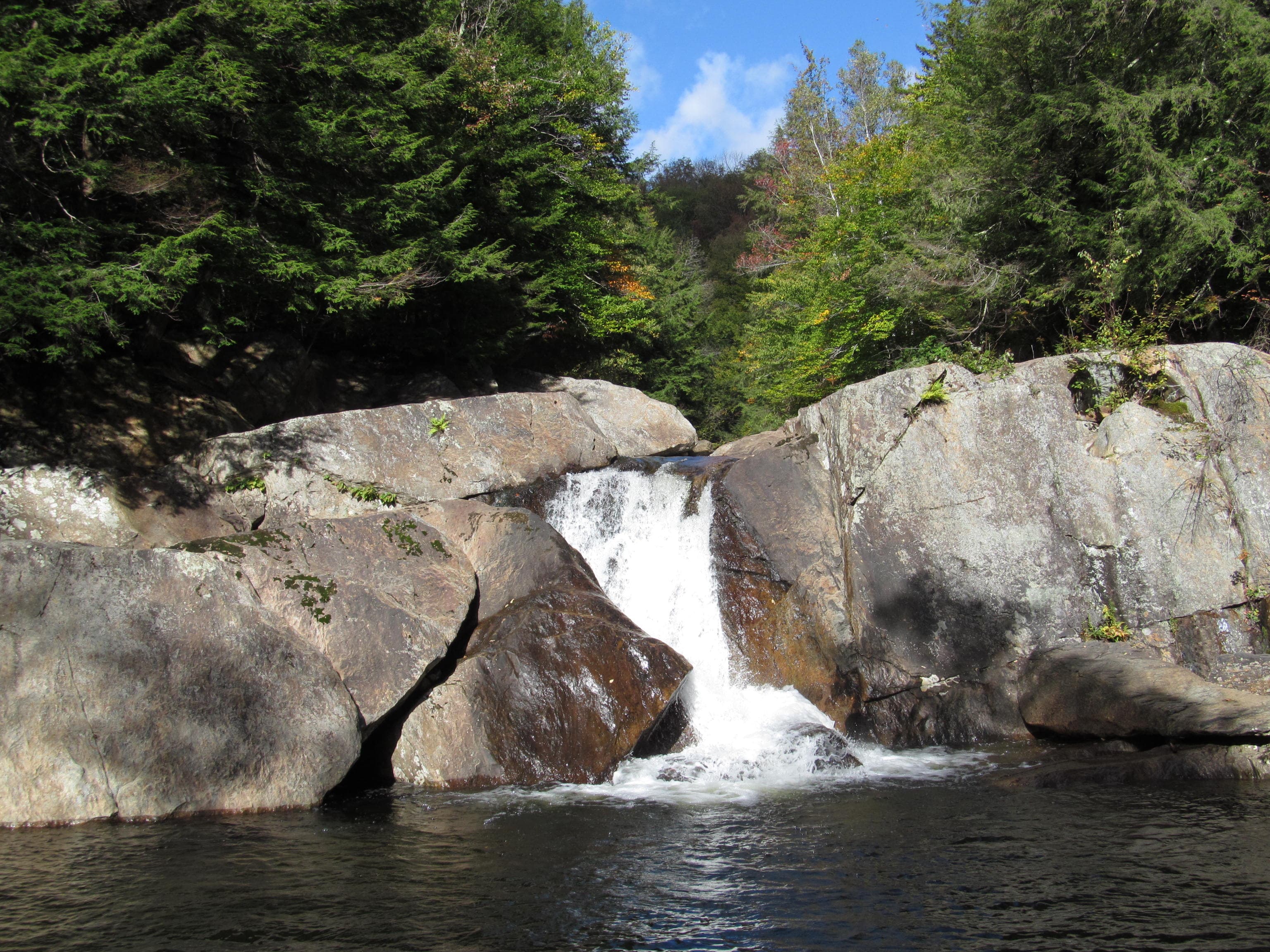 Wide waterfall over grey rock into a dark swimming hole, evergreen and deciduous forest at Buttermilk Falls, Ludlow, Vermont.