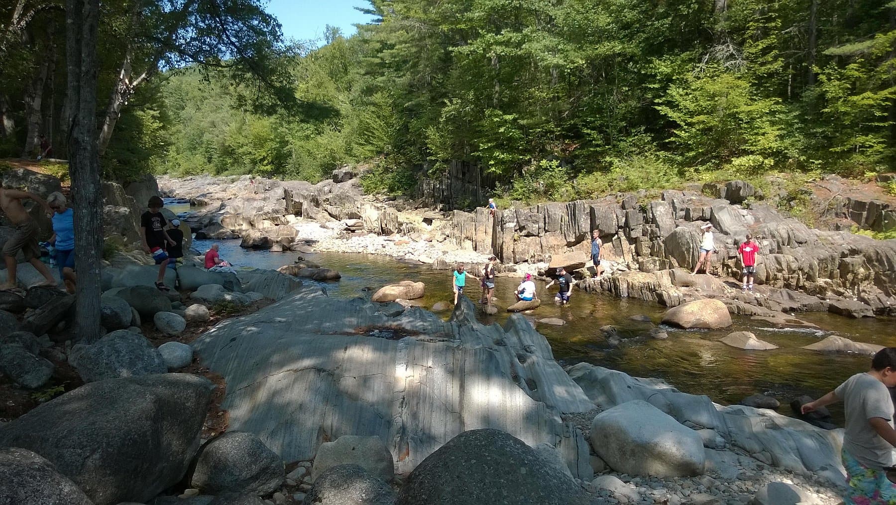 People wading and exploring a broad rocky river gorge in Maine on a summer day.