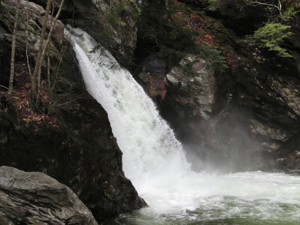 A powerful white waterfall at Bingham Falls cascading over wet, dark rock ledges into frothy water below, framed by evergreens.
