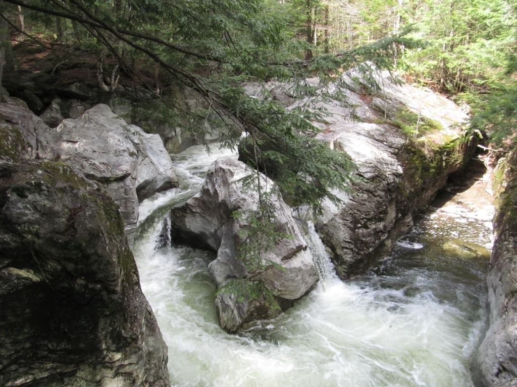 Rushing white water splitting around boulders in a rocky gorge at Bingham Falls, with hemlock branches overhead.
