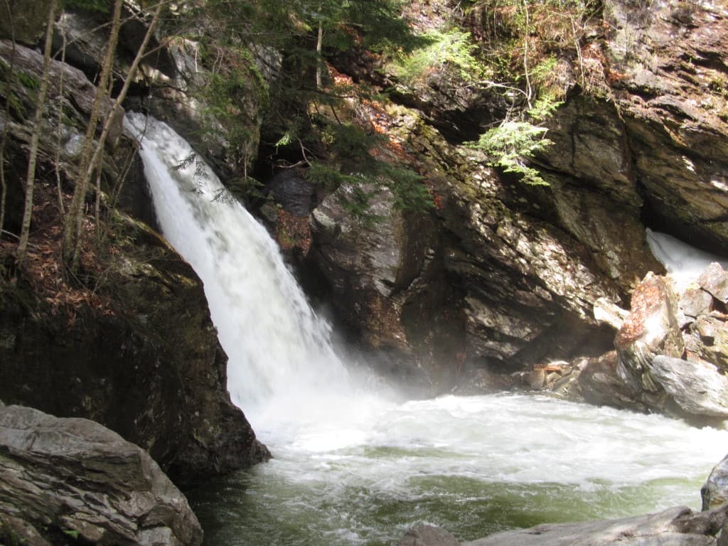 A powerful white waterfall cascading over dark, jagged rock into a deep green pool at Bingham Falls, Vermont.