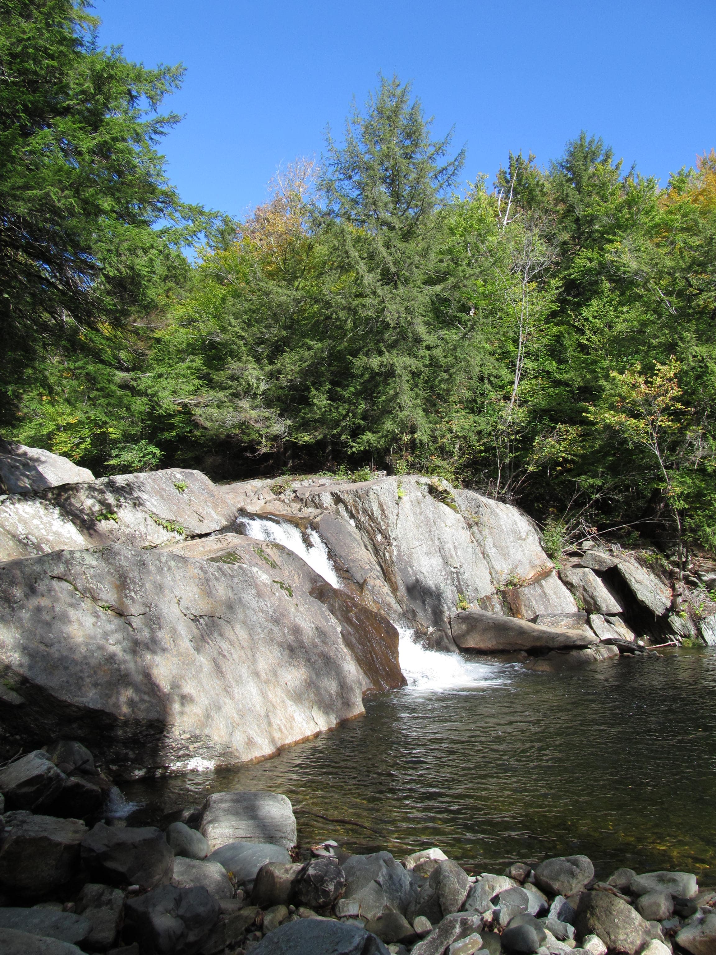 Waterfall cascading over wide grey ledges into a dark pool with a cobble shoreline and forest behind at Buttermilk Falls, Ludlow, Vermont.