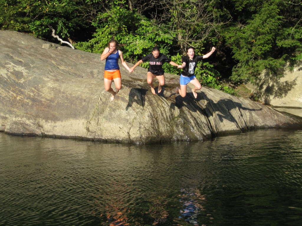 Swimmers shoulder-deep in clear green water at Lareau Swim Hole with a large rock face and summer forest behind them.